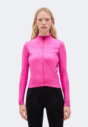 Une femme porte une veste de cyclisme rose vif à fermeture éclair et un cuissard noir sur un fond clair et uni.