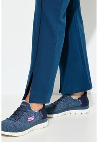 Sportschuhe in Marineblau mit strukturiertem Obermaterial, pinken Akzenten und einer dicken weißen Sohle. Kombiniert mit blauen Hosen mit seitlichen Schlitzen.
