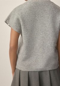 Vue arrière d'une personne portant un pull en tricot à manches courtes gris clair et une jupe plissée grise, sur fond uni.