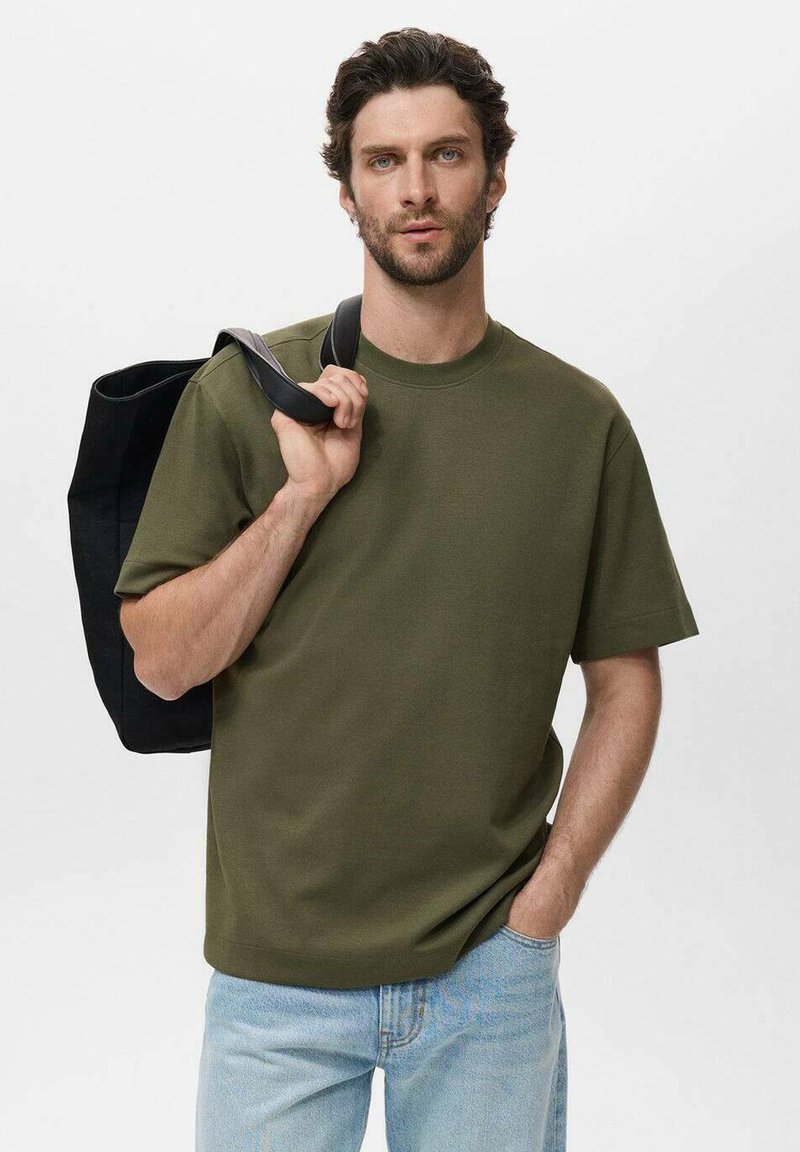 Olive green t-shirt with a crew neck and short sleeves, paired with light blue jeans. The model carries a black tote bag.