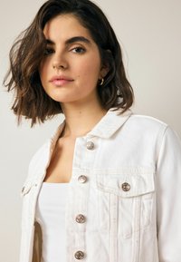 Chaqueta de denim blanca con cierre de botones en la parte delantera, dos bolsillos en el pecho y acentos de metal plateado. La tela parece suave y tiene un corte estructurado.