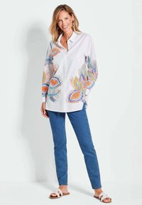 Chemise blanche à boutons avec des broderies florales colorées sur les manches, associée à un jean bleu et des sandales blanches.