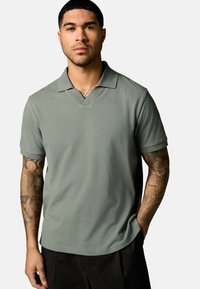 Kortærmet polo-shirt i dæmpet grøn, med en V-hals, blød tekstur og en afslappet pasform. Modellen viser arm-tatoveringer.