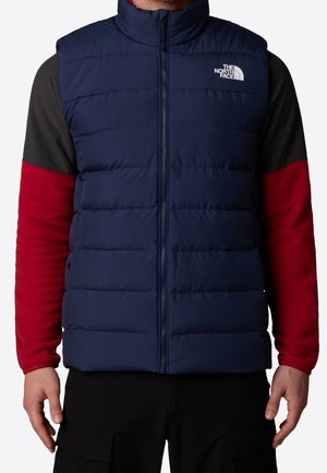Mężczyzna noszący granatową puchową kamizelkę z frontowym suwakiem i logo The North Face na piersi, na długim czarno-czerwonym rękawie koszuli.