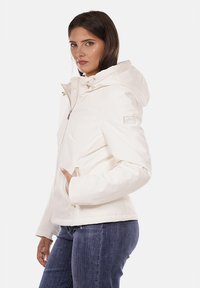 Giacca bianca isolante con cappuccio, zip frontale e tasche. Presenta una texture liscia e accessori in tonificazione oro.