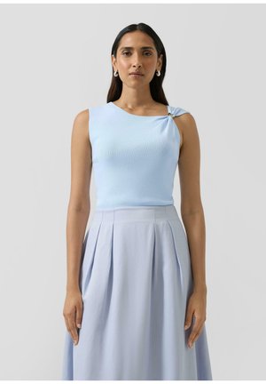 Kobieta o ciemnych włosach w jasnoniebieskim topie bez rękawów z ozdobą na ramieniu oraz dopasowanej plisowanej spódnicy, stojąca na tle szarego tła.