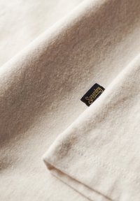 Tissu doux couleur crème avec une texture tricotée visible et une petite étiquette noire portant l'inscription "Superdry" cousue près de l'ourlet.