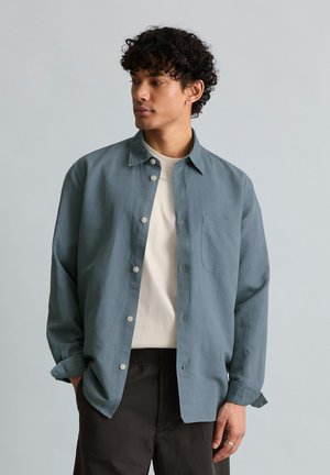 Junger Mann mit lockigem Haar, der ein blaues Hemd über einem weißen Oberteil und schwarze Hosen trägt, schaut seitlich vor einem schlichten Hintergrund.