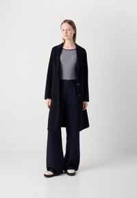 Manteau en laine bleu marine avec une coupe ajustée, deux boutons, associé à un haut rayé à manches longues et un pantalon large bleu marine. Chaussures à enfiler de couleur claire.