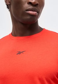 Κοντινό πλάνο ενός ατόμου που φοράει φωτεινό πορτοκαλί μπλουζάκι με στρογγυλή λαιμόκοψη και μικρό μαύρο λογότυπο Reebok στο στήθος.