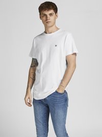 Weißes Baumwoll-T-Shirt mit Rundhalsausschnitt und kurzen Ärmeln, mit einem kleinen schwarzen Logo auf der linken Brust. Kombiniert mit blauen Jeans.