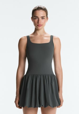 Frau mit zurückgebundenem Haar, die ein ärmelloses dunkelgraues eng anliegendes Kleid mit einem gerafften, ballonartigen Rock trägt, steht vor einem schlichten Hintergrund.