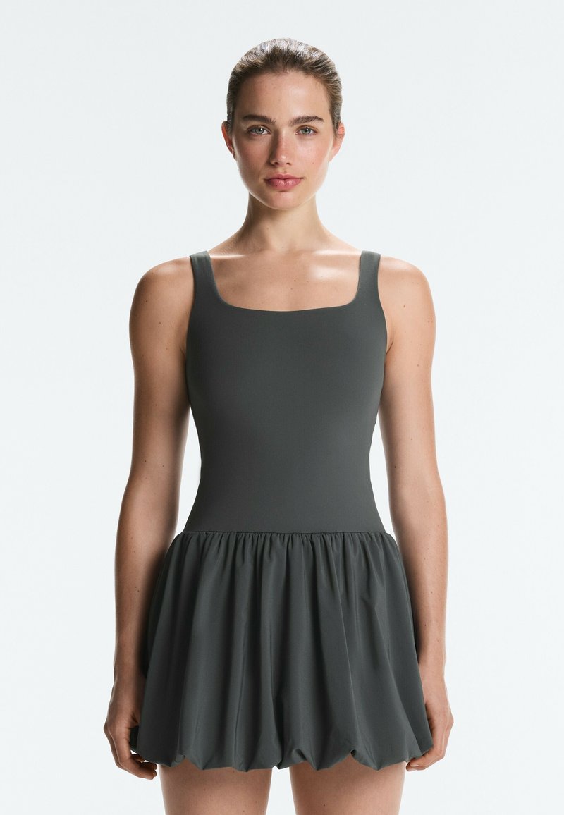 Femme aux cheveux tirés en arrière portant une robe ajustée sans manches gris foncé avec une jupe bouffante froncée, debout devant un fond uni.