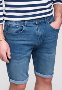 Mann trägt blaue hochgekrempelte Denim-Shorts und ein blau-weiß horizontal gestreiftes Langarmshirt, mit einer Hand in der Tasche.