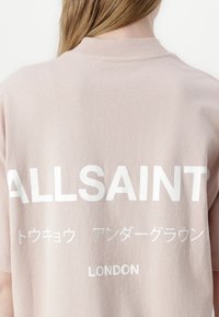 Lys rosa bomull T-skjorte med fet hvit tekst på baksiden, inkludert "ALLSAINTS" og "LONDON," med rund hals og korte ermer.