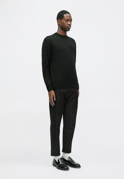 Vyras stovi apsivilkęs juodą megztinį, juodas trumpas kelnes, baltas kojines ir juodus loaferius prieš paprastą baltą foną.