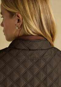 Brūns ķīmis joprojām jaka ar ribotu apkakli un izšūtu "JOULES" logo. Gluda tekstūra ar dimanta rakstu detaļām.