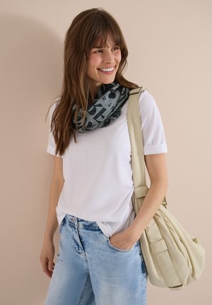 Femme souriante portant un t-shirt blanc, un jean bleu clair, une écharpe noire à motifs et un grand sac à bandoulière beige sur un fond uni.