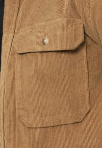 Chemise en velours côtelé de couleur beige chaud, avec une texture côtelée verticale, une poche à rabat et un col arrondi avec un bouton-pression en métal.