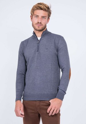 Jeune homme aux cheveux blonds portant un pull gris à fermeture éclair au col avec des coudières et un pantalon marron, debout devant un fond clair.