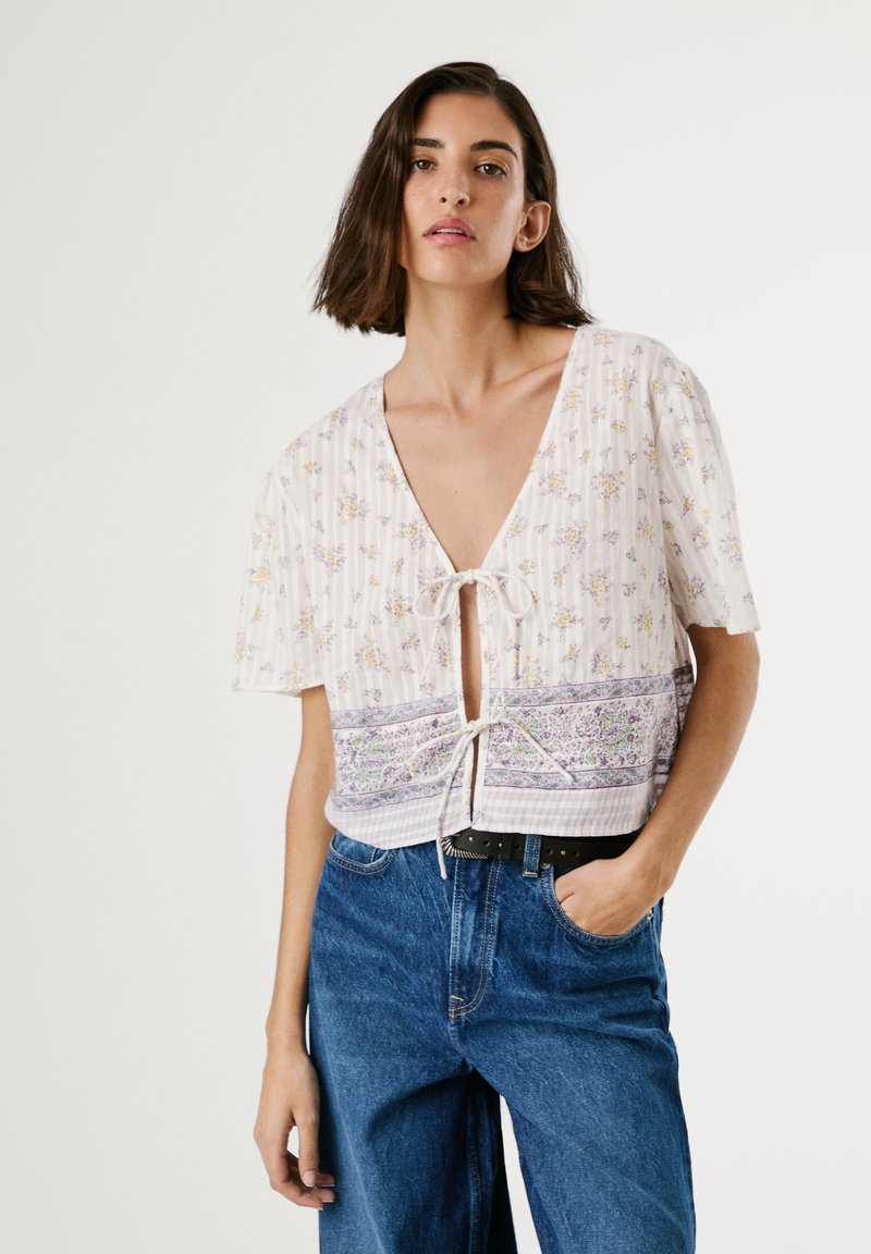 Kortärmad blus med blommönster i lätt material och knytframsida, prydd med lila och gula blommor på vit bakgrund, lager på lager med ett mönstrat band.