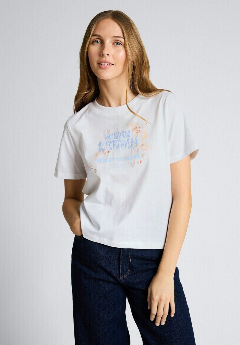 Mujer joven con camiseta blanca con gráficos en azul y naranja y vaqueros azul oscuro, de pie frente a un fondo claro y liso.