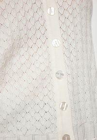 Cardigan bianco lavorato a maglia con motivo a rombi, dotato di una chiusura con sei bottoni rotondi e trasparenti, e finiture a coste all'orlo e ai polsini.