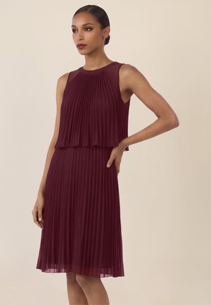 Kobieta z włosami spiętymi w kok, ubrana w bezrękawową, plisowaną sukienkę koloru bordowego, stoi z jedną ręką na biodrze na beżowym tle.