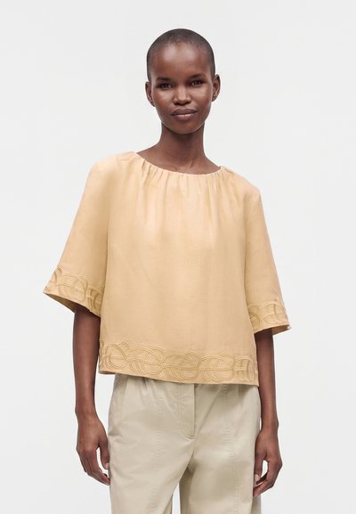 Femme portant un chemisier beige ample avec un ourlet et des bords de manches brodés, associé à un pantalon beige clair, debout devant un fond uni.