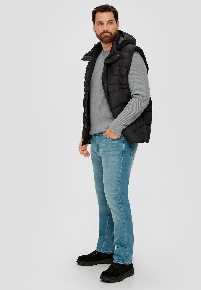 Black padded vest over a gray sweater, light blue jeans, and black footwear. The vest features a hood and zip closure.