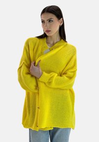 Żółty sweter na drutach z fakturą, o luźnym kroju, zaokrąglonym dekoltem i zapięciem na guziki z przodu, stylizowany z jasnoniebieskimi dżinsami.