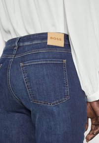 Tmavě modré denimové džíny mají na pasu béžovou koženou nášivku s embosovaným nápisem "BOSS". Džíny mají standardní zadní kapsy a viditelné švy.