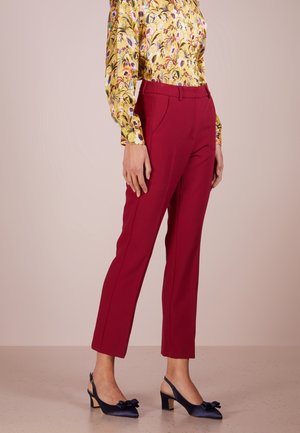 Frau trägt rote, maßgeschneiderte Hose, gelbe langärmelige Bluse mit floralem Muster und marineblaue Slingback-Absätze mit Schleifen, stehend vor einem einfarbigen Hintergrund.