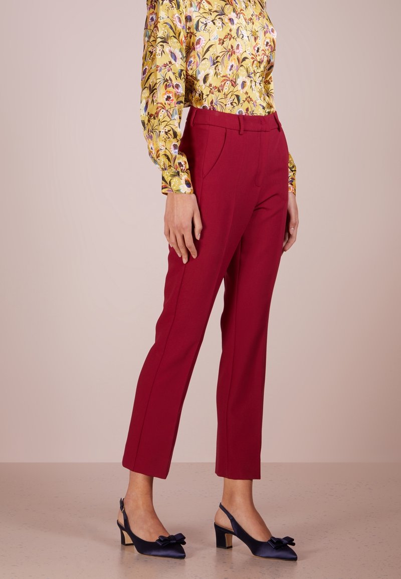 Femme portant un pantalon rouge sur mesure, un chemisier jaune à manches longues avec motifs floraux et des talons slingback bleu marine avec des nœuds, debout devant un fond uni.