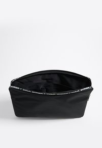 Bolsa de tela negra con una abertura amplia, que cuenta con un cierre de cremallera y un bolsillo interior. Decorada con una correa con logotipo a lo largo del borde superior.
