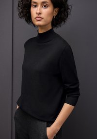 Pull en maille noir avec un col montant, des manches longues et une coupe décontractée. Texture lisse, associé à un pantalon gris foncé.