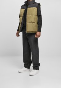 Pufferväst med svart nylontyg på ovansidan och olivgrön vaddering på undersidan, med ståkrage och dragkedja. Matchas med svarta jeans och vita sneakers.