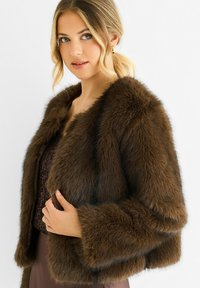 Young woman with blonde hair in a low ponytail wearing gold hoop earrings and a dark brown faux fur jacket over a textured top.