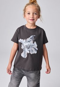 Mladé dieťa so svetlými vlasmi v cope, oblečené v čiernom tričku AllSaints s bielym kvetinovým vzorom a sivých džínsoch, stojace pred jednofarebným pozadím.