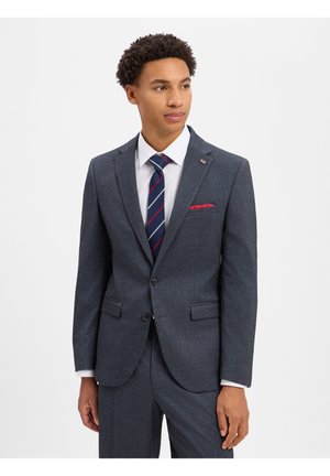Giovane uomo che indossa un abito grigio scuro con camicia bianca, cravatta a righe blu e rosse e fazzoletto rosso nel taschino, guardando leggermente di lato.