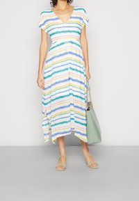 Femme portant une robe à rayures multicolores avec un col en V, portant un sac à main vert clair et des sandales assorties à brides.