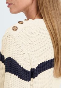 Kremfarget strikket genser med en marineblå horisontal stripe, med tre brune knapper på skulderen og et teksturert, åpent vevdesign.