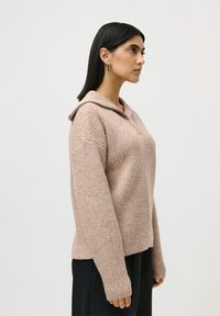 Kvinne med rett svart hår iført hoopsmykker og en beige glidelåsstrikkegenser som står i profil mot en hvit bakgrunn.