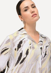 Blusa con colletto dal profilo fluido in bianco, caratterizzata da strisce astratte in nero, grigio e giallo. Tessuto liscio con una leggera lucentezza.