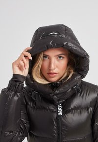 Mujer rubia ajustando la capucha de una chaqueta de plumas negra brillante con etiquetas de marca en la capucha y el zipper, contra un fondo sencillo.