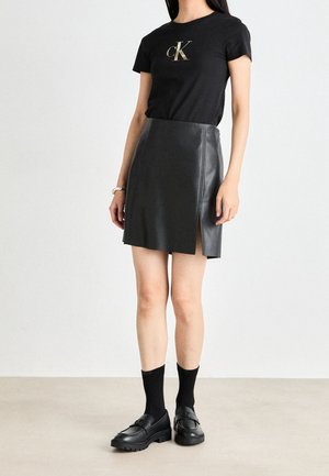 Mujer que lleva una camiseta negra Calvin Klein, minifalda de cuero negra con abertura lateral, calcetines negros y mocasines negros.