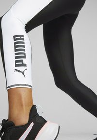 Svartvita leggings med en framträdande Puma-logotyp och kattgrafik, kombinerade med svarta sportskor med rosa detaljer och skosnören.