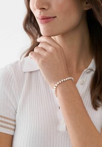 Armbandet i guldton med vita cirkulära pärlor bärs på en handled. Personen är klädd i en ribbad vit polo med krage.