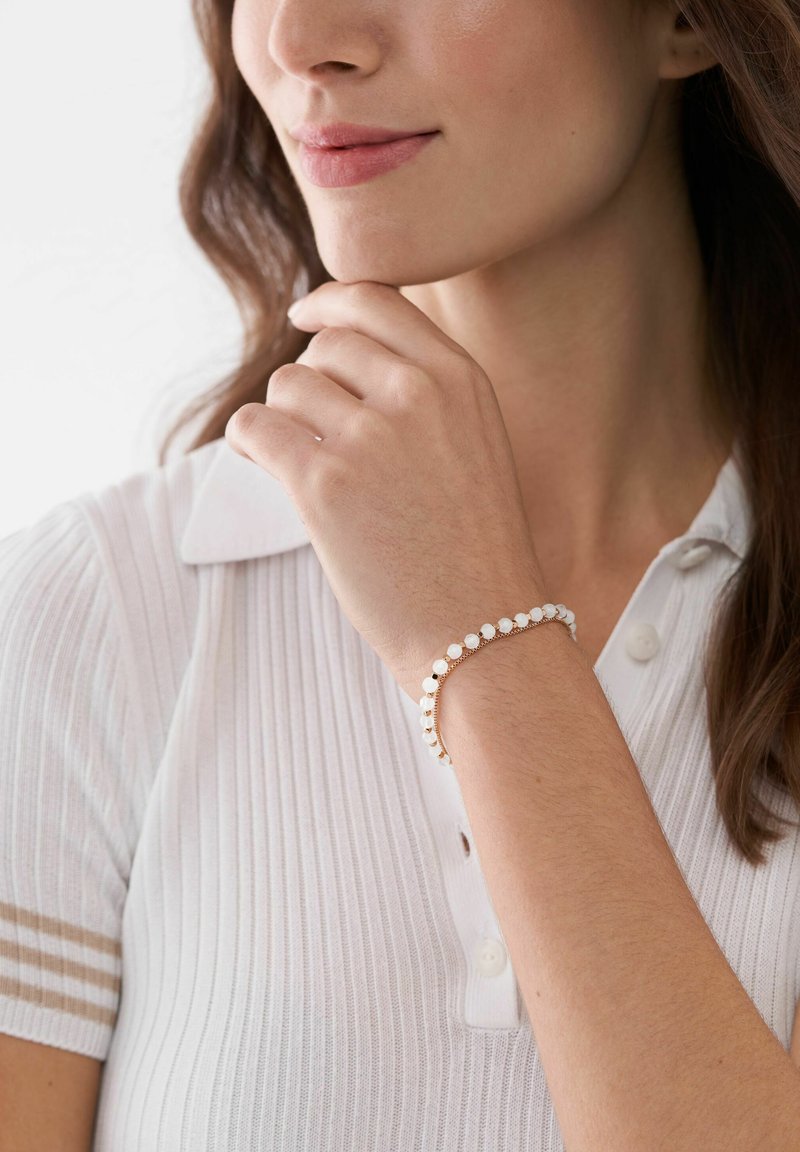 Armbandet i guldton med vita cirkulära pärlor bärs på en handled. Personen är klädd i en ribbad vit polo med krage.
