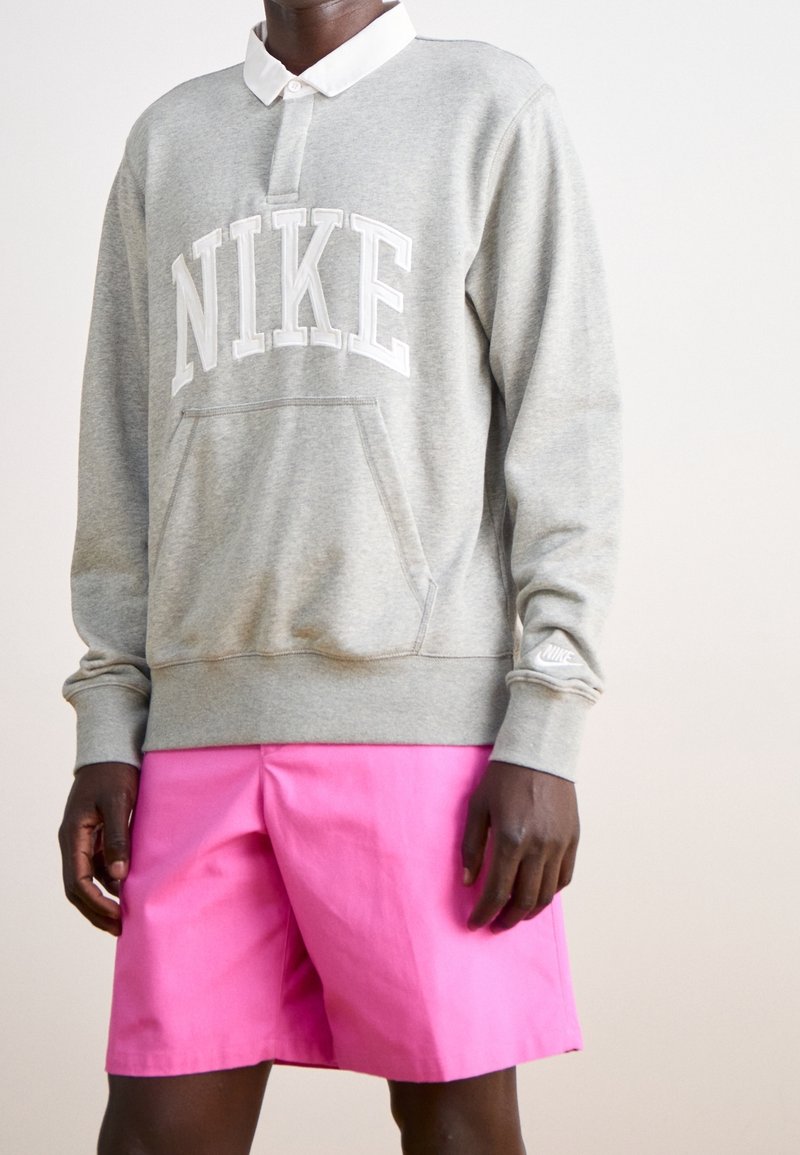 Szara bluza z napisem "NIKE" w białej czcionce, z białym kołnierzykiem i kieszenią z przodu, w połączeniu z jaskrawo różowymi szortami.